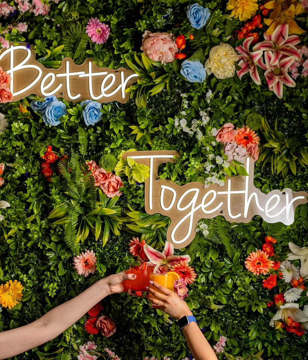 Two people clinking colorful drinks in front of a floral wall at Effy’s Café